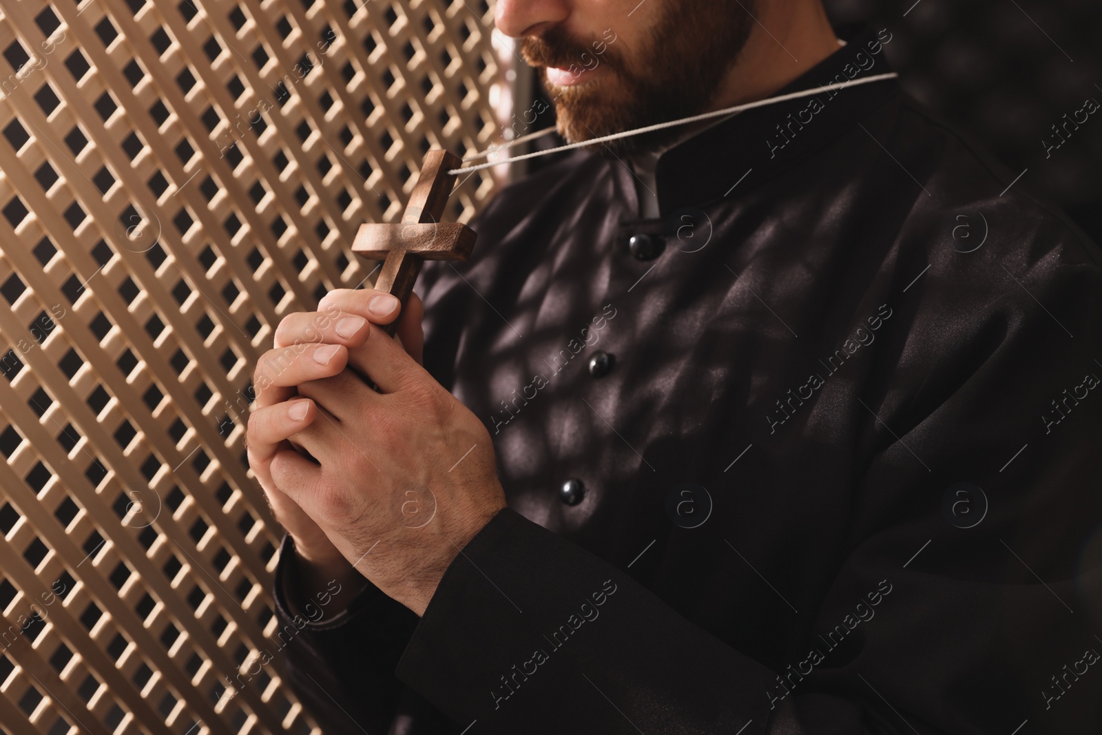 Photo of Catholic priest in cassock holding cross in confessional booth, closeup