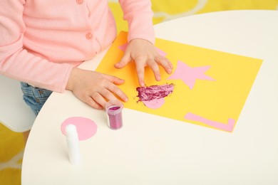 Girl using colorful glitter while making paper card at desk in room, closeup. Home workplace