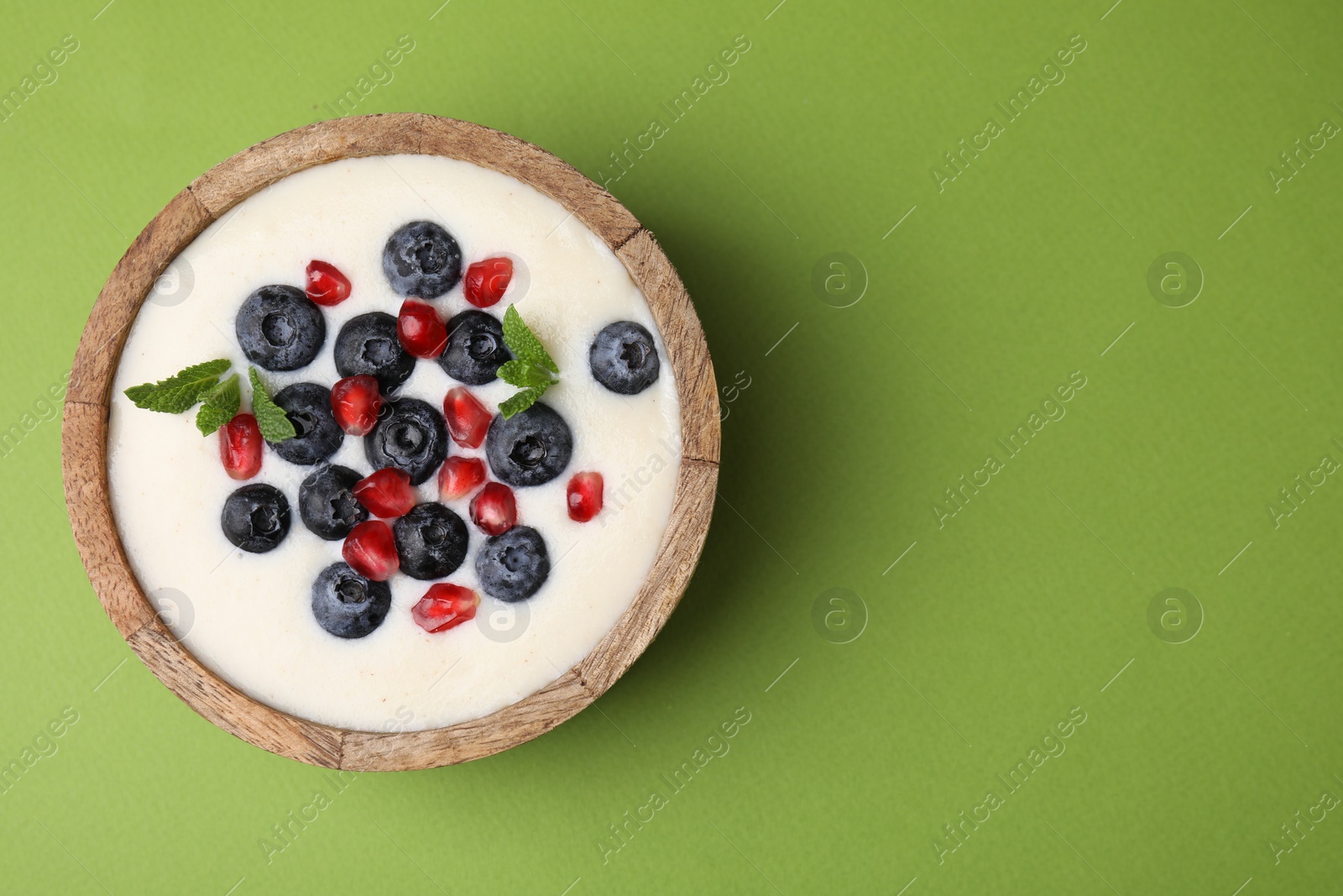 Photo of Bowl of delicious semolina pudding with blueberries, pomegranate and mint on green background, top view. Space for text