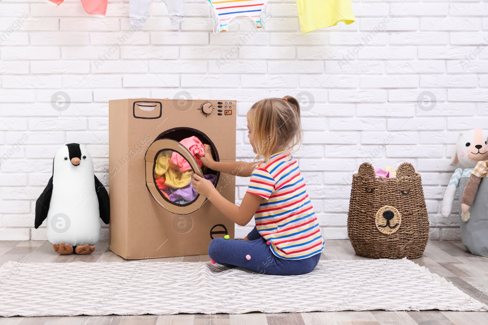 Photo of Little girl putting laundry into toy cardboard washing machine indoors