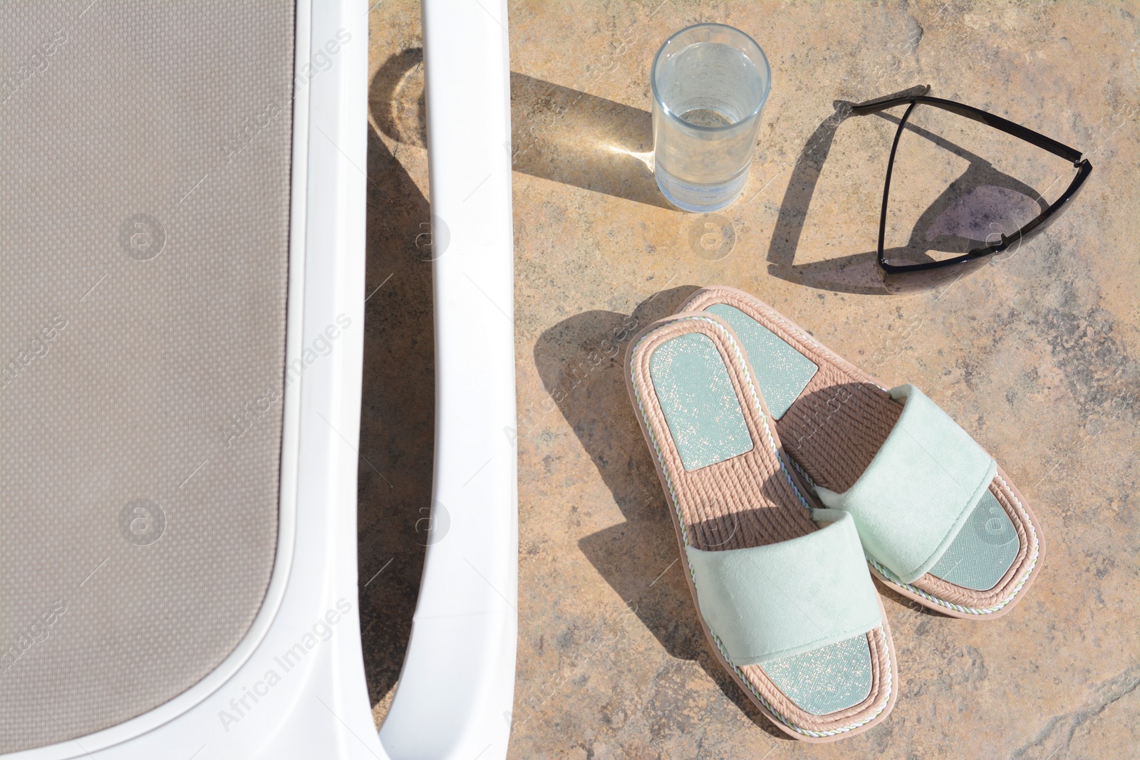 Photo of Stylish slippers, sunglasses, glass of water and grey sunbed outdoors. Beach accessories