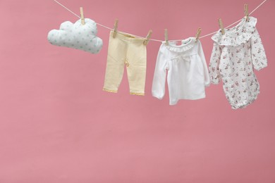 Photo of Different baby clothes and cloud shaped pillow drying on laundry line against pink background. Space for text