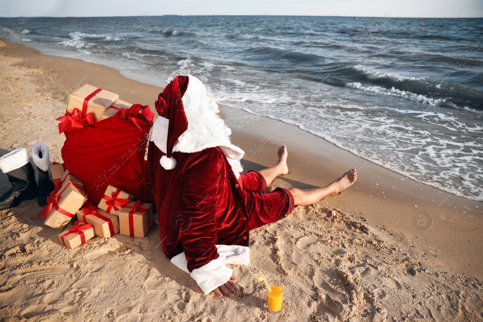 Photo of Santa Claus with cocktail and bag of presents relaxing on beach. Christmas vacation