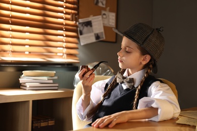 Photo of Cute little detective with smoking pipe at table in office. Space for text