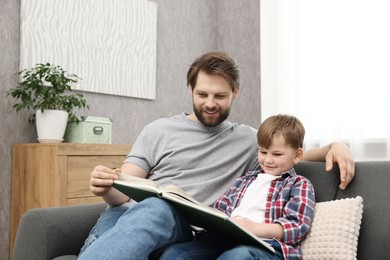 Photo of Happy dad and son reading book together on sofa at home