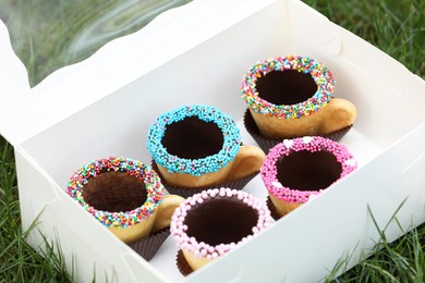 Photo of Box of delicious edible biscuit coffee cups decorated with sprinkles on green grass outdoors, closeup