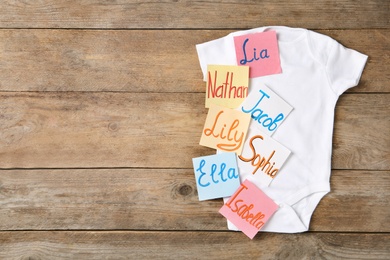 Paper notes with different baby names and child's bodysuit on wooden table, top view. Space for text