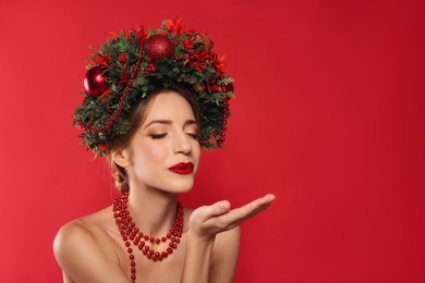 Photo of Beautiful young woman with Christmas wreath blowing kiss on red background. Space for text