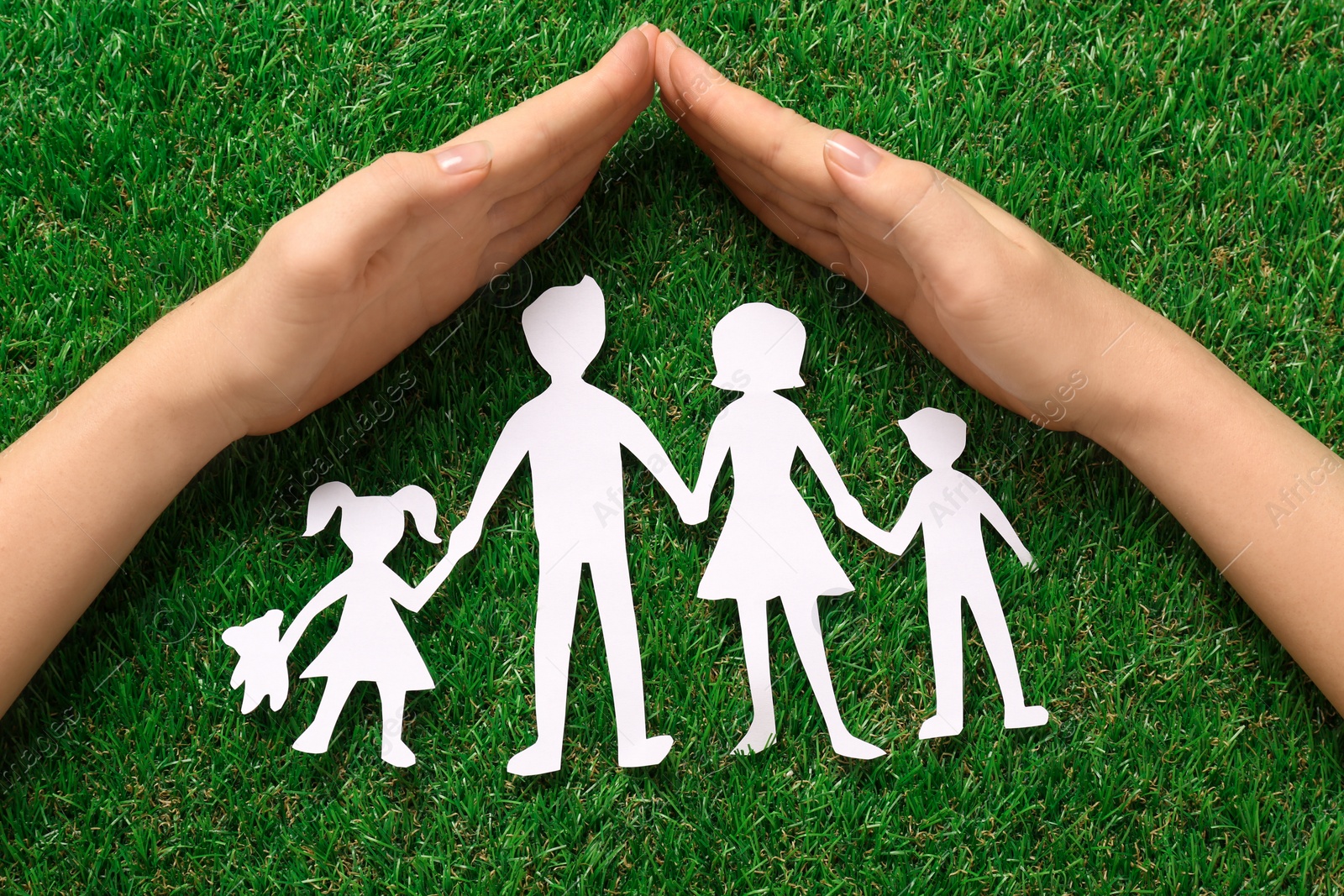 Photo of Woman covering family paper cutout with hands on green grass, top view