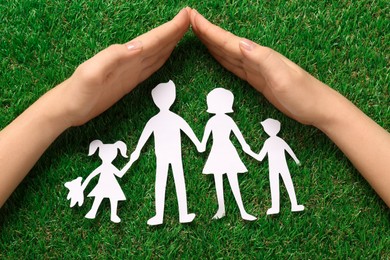 Woman covering family paper cutout with hands on green grass, top view