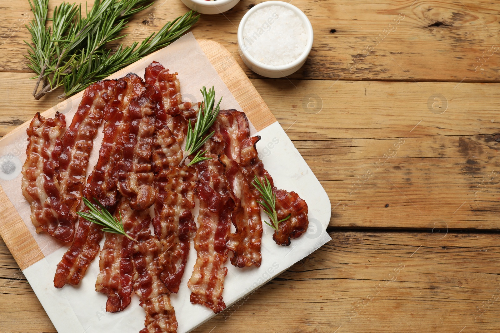 Photo of Slices of tasty fried bacon, rosemary and salt on wooden table, top view, space for text