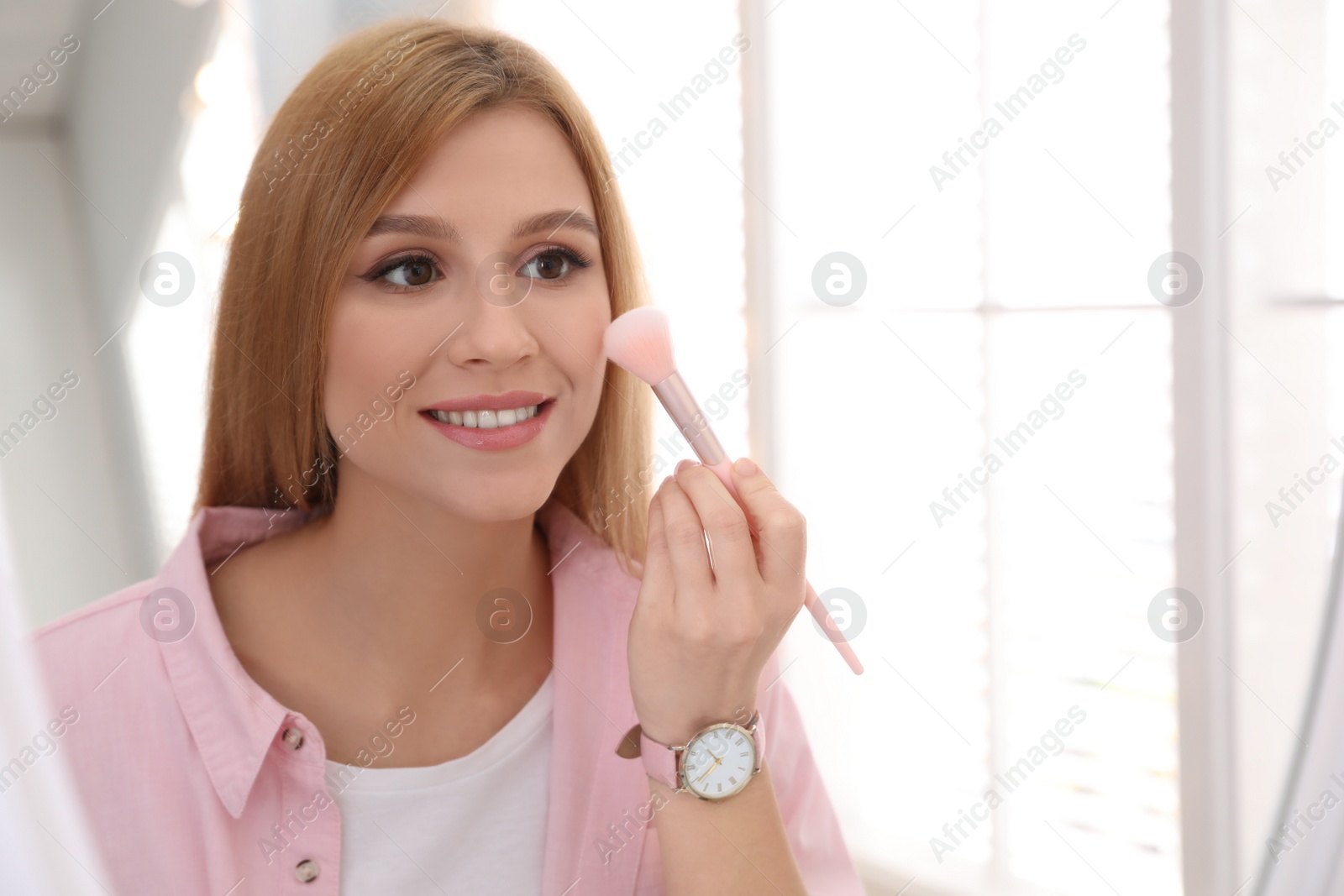 Photo of Beautiful young woman applying makeup near mirror indoors