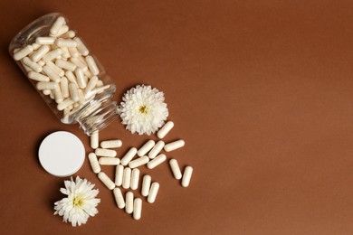 Open medicine bottle, scattered pills and flowers on brown background, flat lay. Space for text