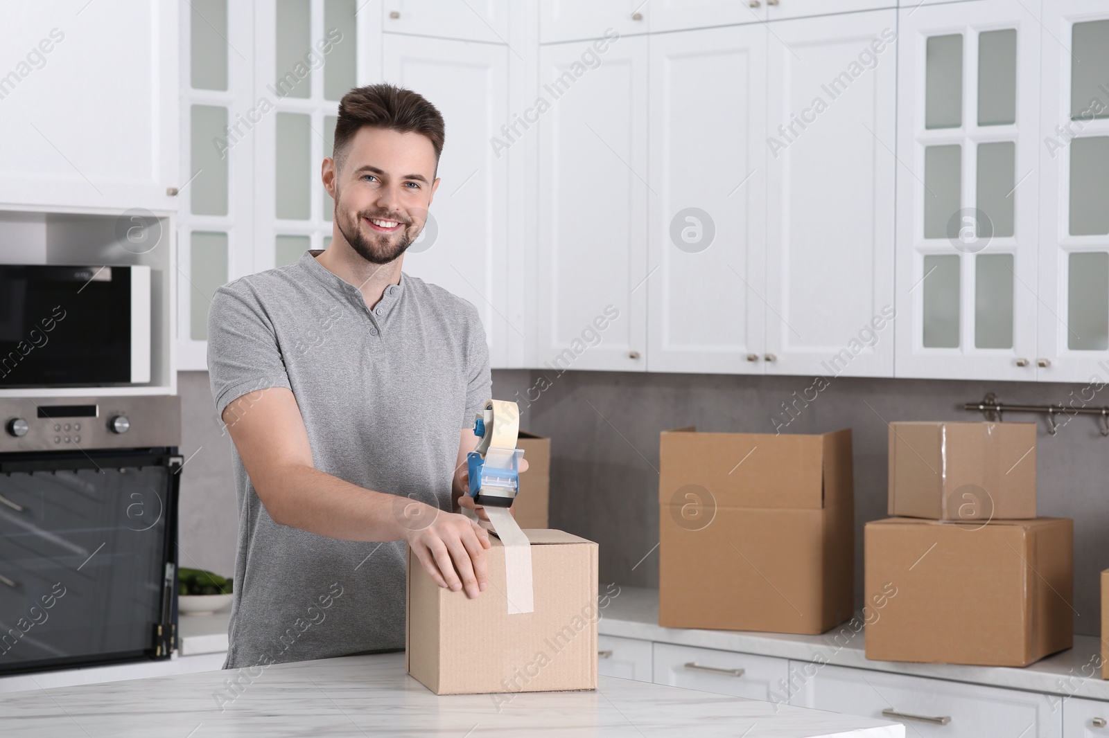 Photo of Smiling man taping box with adhesive tape dispenser in kitchen, space for text