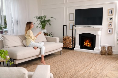 Photo of Young woman watching television at home. Living room interior with TV on fireplace