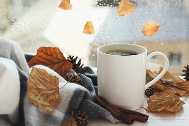 Photo of Cup of hot drink and autumn leaves near window on rainy day. Cozy atmosphere