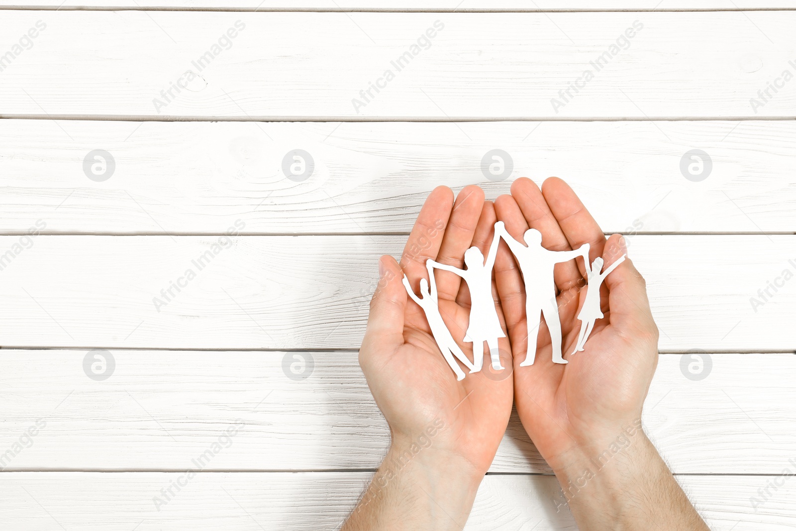 Photo of Young man holding paper family figure in his hands against white wooden background, top view. Space for text