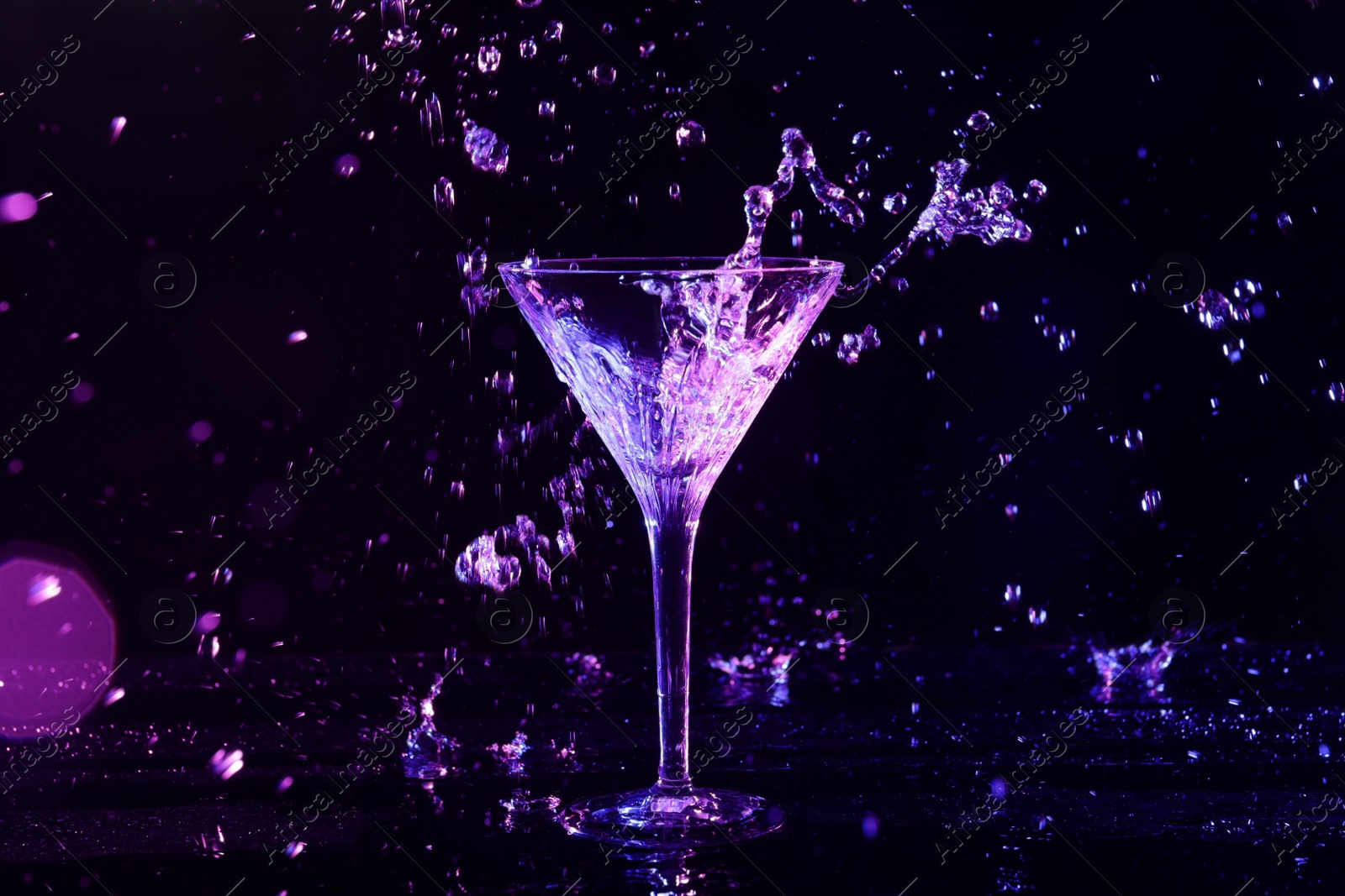 Photo of Glass of martini and splashes in neon lights on dark background, bokeh effect