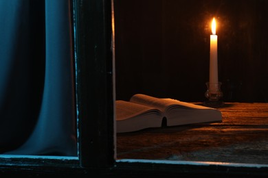 Photo of Burning candle and Bible on wooden table at night, view through window