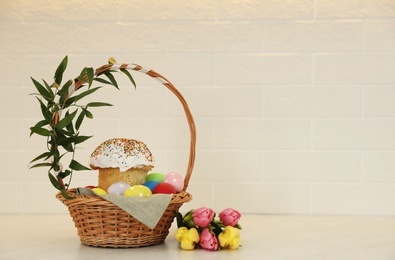 Basket with traditional Easter cake, dyed eggs and flowers on table near white brick wall. Space for text