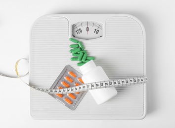 Weight loss pills, bottle, scales and measuring tape on white background, top view