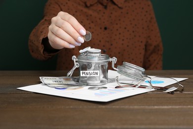 Photo of Pension concept. Woman putting coin into glass jar with money at wooden table, closeup