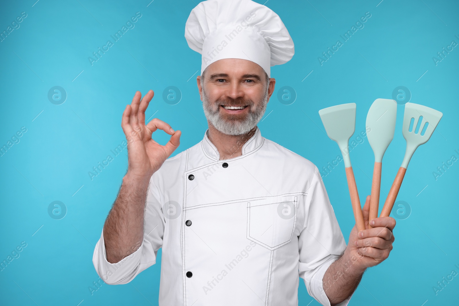 Photo of Happy chef in uniform with kitchen utensils showing OK gesture on light blue background