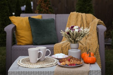 Photo of Cups of drink, cheese, croissants and autumn decor on table outdoors. Garden rattan furniture