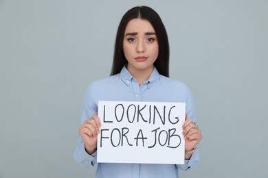 Photo of Young unemployed woman holding sign with phrase Looking For A Job on grey background