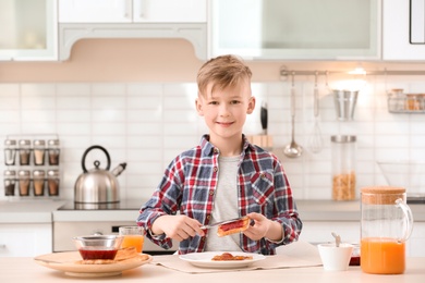 Photo of Cute little boy spreading jam onto tasty toasted bread at table in kitchen