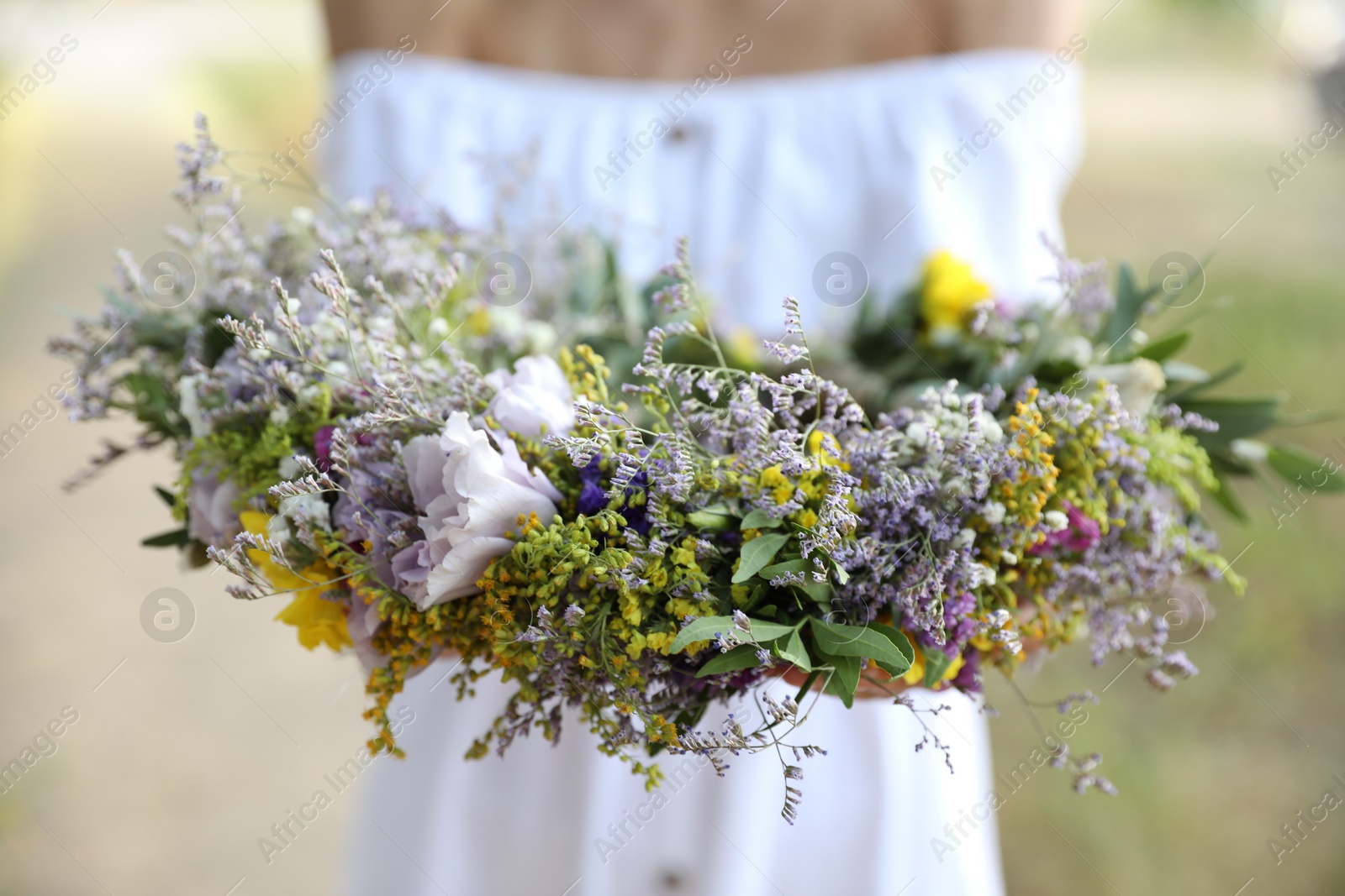 Photo of Young woman holding wreath made of beautiful flowers outdoors on sunny day, closeup