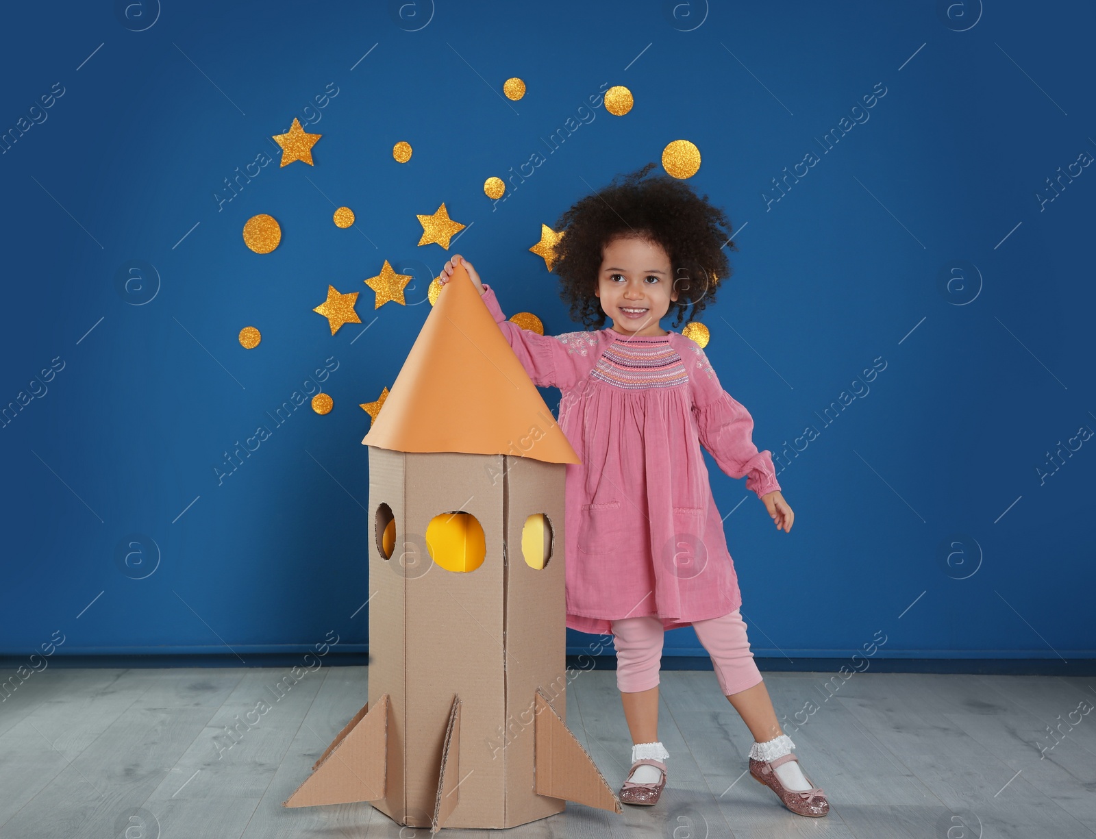 Photo of Cute African American child playing with cardboard rocket near blue wall