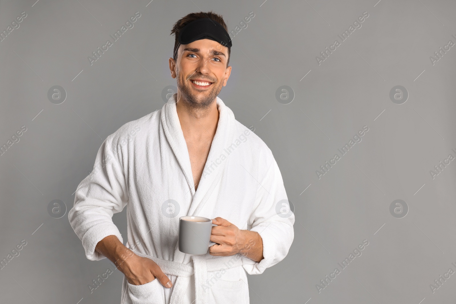 Photo of Happy young man in bathrobe with cup of coffee on grey background, space for text