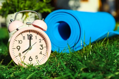 Photo of Alarm clock and fitness mat on green grass outdoors, closeup with space for text. Morning exercise