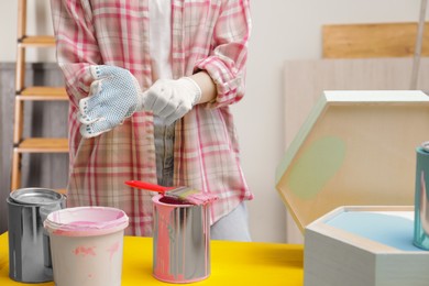Photo of Woman wearing gloves near paint cans and wooden shelf at yellow table indoors, closeup
