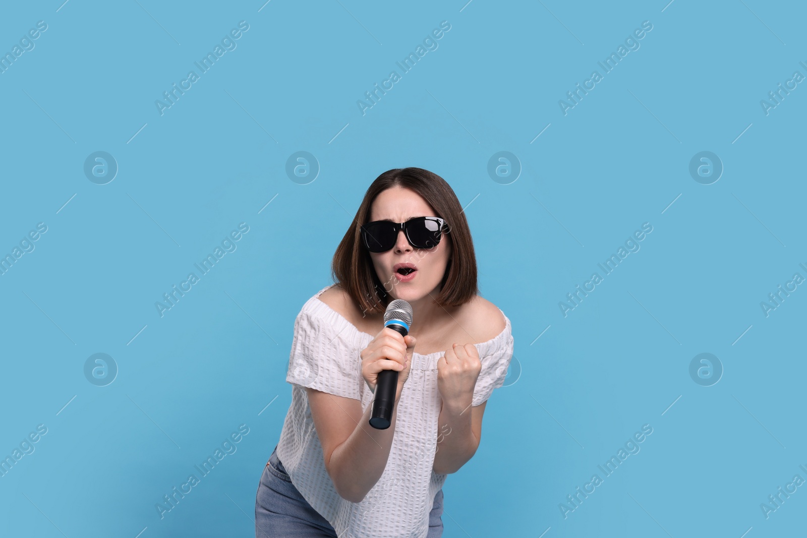 Photo of Beautiful young woman with sunglasses and microphone singing on light blue background