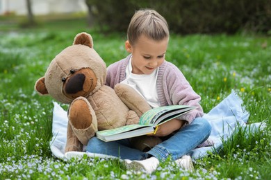 Little girl with teddy bear and book on plaid outdoors