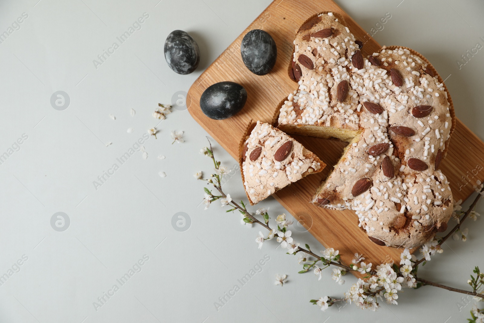 Photo of Board with cut delicious Italian Easter dove cake (traditional Colomba di Pasqua), painted eggs and flowering branches on light grey table, flat lay. Space for text