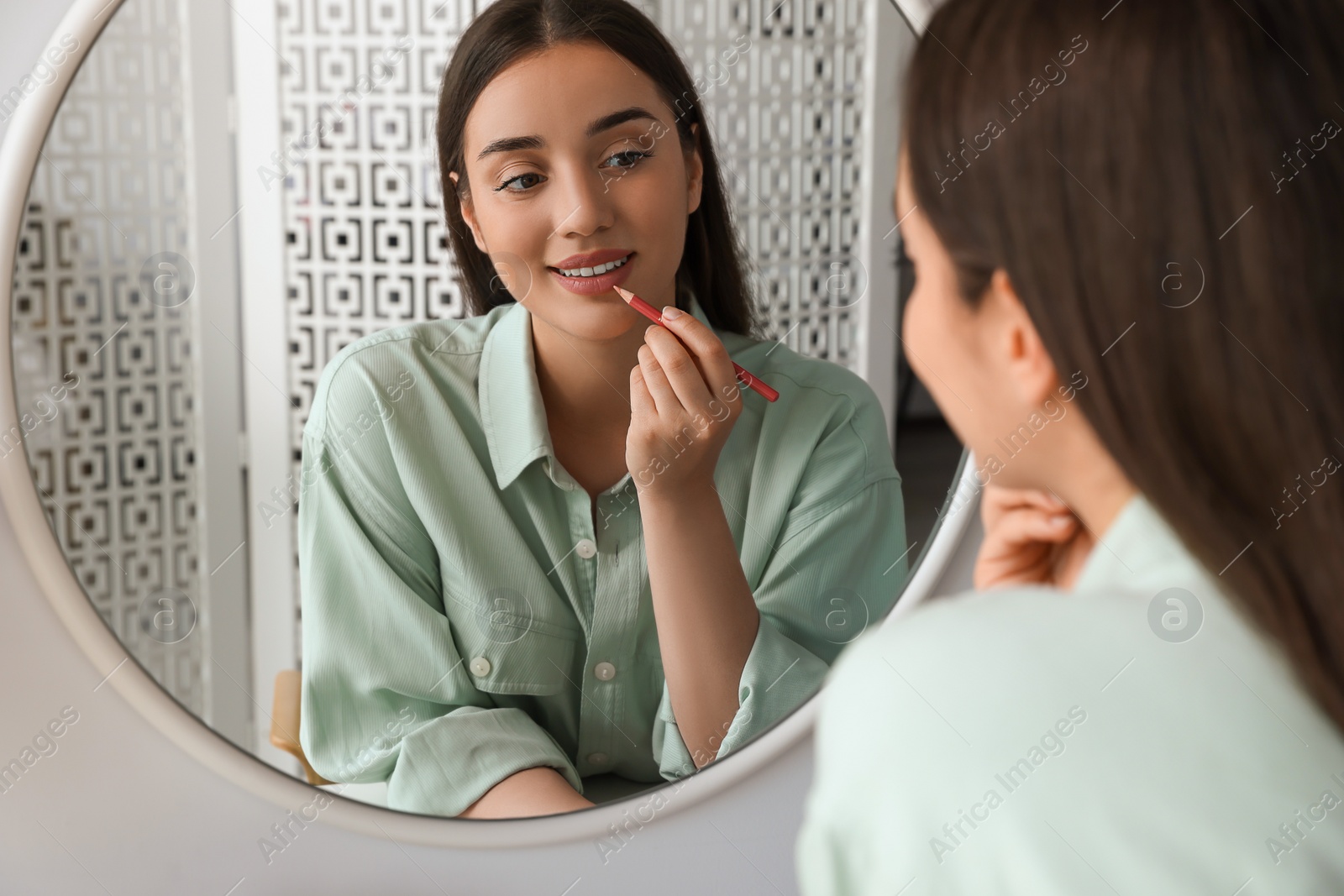 Photo of Beautiful young woman applying cosmetic pencil on lips near mirror indoors