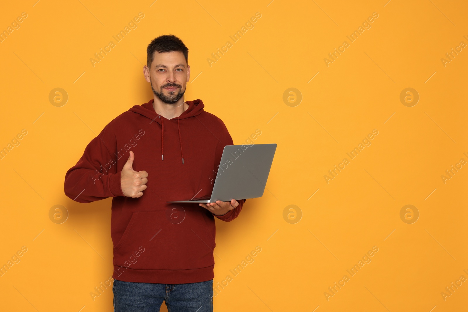 Photo of Handsome man with laptop showing thumbs up on orange background. Space for text