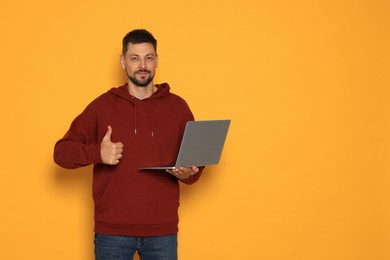 Photo of Handsome man with laptop showing thumbs up on orange background. Space for text