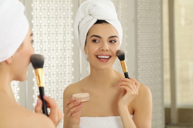 Beautiful young woman applying face powder with brush in front of mirror at home