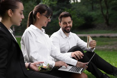 Photo of Happy colleagues with laptop having business lunch on green grass outdoors, selective focus