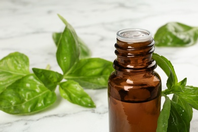 Bottle of basil essential oil and fresh leaves on marble table, closeup. Space for text