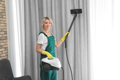 Photo of Female janitor removing dust from curtain with steam cleaner indoors