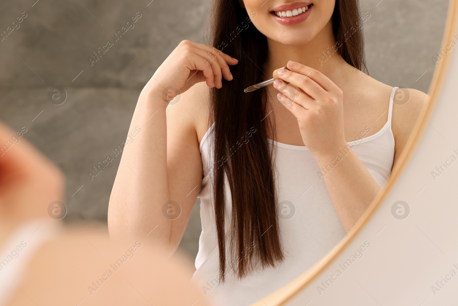 Photo of Happy woman applying essential oil onto hair near mirror, closeup