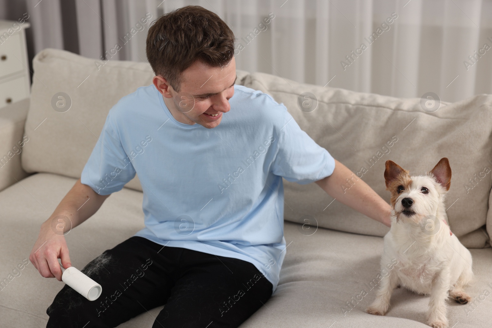 Photo of Pet shedding. Smiling man with lint roller removing dog's hair from pants on sofa at home