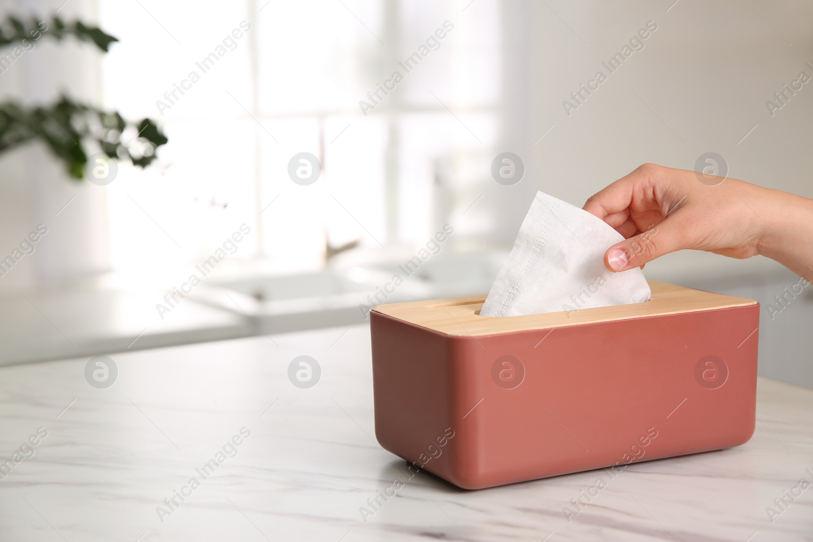 Photo of Woman taking paper tissue out of box on white marble table in kitchen, closeup. Space for text