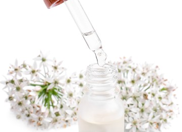 Photo of Dripping essential oil from pipette into bottle near garlic chives flowers on white background, closeup