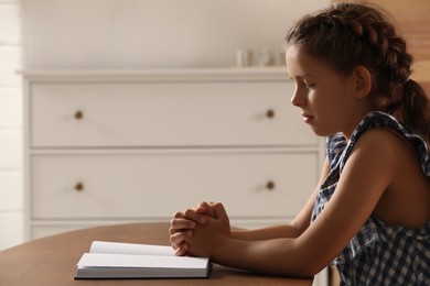 Cute little girl praying over Bible at table in room